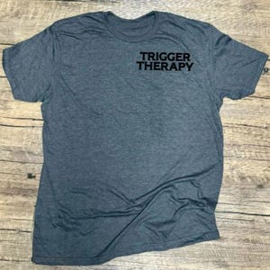 May include: A heather gray crew neck t-shirt with the text "TRIGGER THERAPY" printed in black on the upper chest. The shirt is made of a soft material and is displayed on a wooden surface.