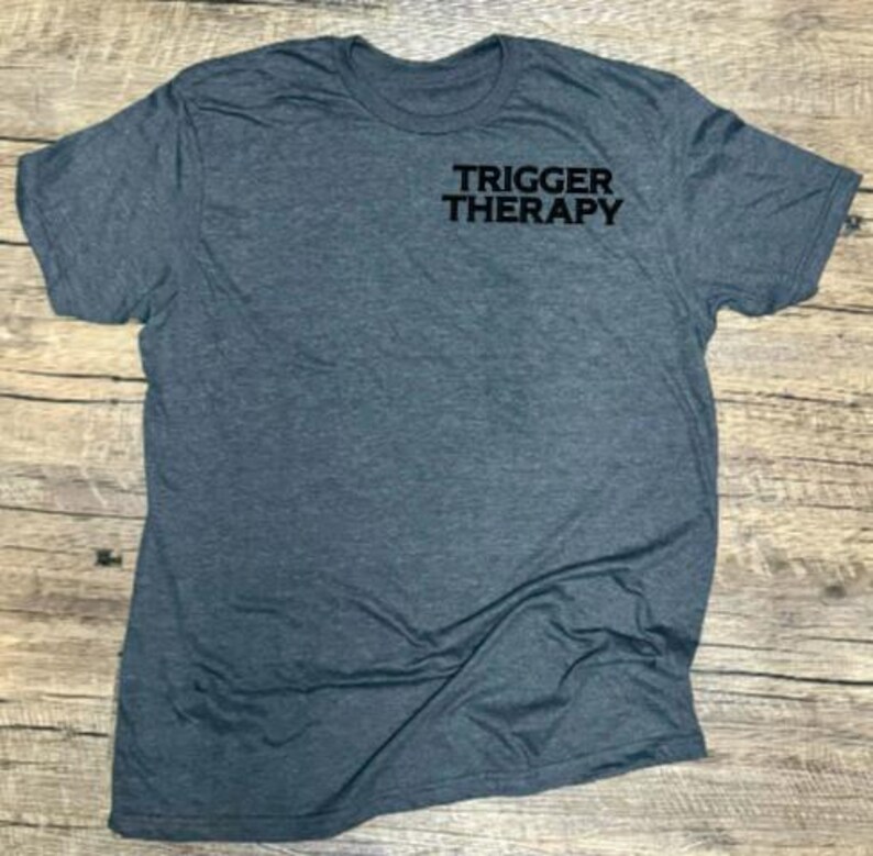 May include: A heather gray crew neck t-shirt with the text "TRIGGER THERAPY" printed in black on the upper chest. The shirt is made of a soft material and is displayed on a wooden surface.
