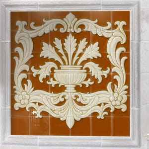 May include: A decorative tile panel featuring a cream-colored, ornate floral design against a warm, reddish-brown background. The design includes a central vase with stylized leaves and scrolling embellishments, framed by a white border. The tile is mounted on a tiled wall.