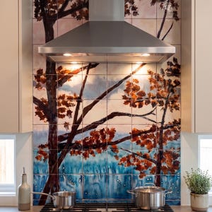 May include: Kitchen backsplash with a hand-painted tree design. The artwork features a tree with brown branches and orange leaves against a blue and white background. A stainless steel range hood is above the backsplash. Two stainless steel pots sit on the gas stove.