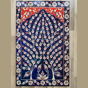May include: A rectangular tile mural featuring a tree design with white flowers and red accents against a deep blue background. The border is adorned with floral patterns. The artwork is displayed on a wooden surface.