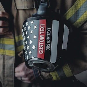 May include: A black helmet band featuring a white star pattern on the left side, a vertical red stripe, and the text CUSTOM TEXT printed in white. The band is attached to a tan firefighter jacket with reflective yellow and grey safety stripes.