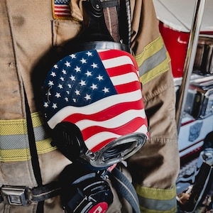 May include: A black firefighter's breathing apparatus mask featuring a prominent American flag graphic on the front, worn over a tan turnout coat with yellow reflective stripes and a small American flag patch on the chest area.
