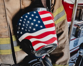May include: A black firefighter's breathing apparatus mask featuring a prominent American flag graphic on the front, worn over a tan turnout coat with yellow reflective stripes and a small American flag patch on the chest area.