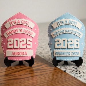 May include: Two decorative baby announcement plaques. One is pink with the text "IT'S A GIRL", "DADDY'S HERO", "2025", and "AURORA". The other is blue with "IT'S A BOY!", "KESTON HATCHETTE", "2026", and "SUMMER 2026".