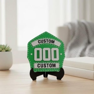 A single 3D printed Fire Helmet Shield in a vibrant green and white color combination, displayed on a black easel stand. The customizable PLA plaque features large white 000 numbers and CUSTOM banners, set on a clean desk with soft lighting, making it an appealing item for fire station office decor or a unique fireman's birthday gift.