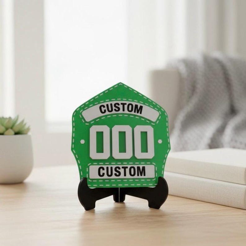 A single 3D printed Fire Helmet Shield in a vibrant green and white color combination, displayed on a black easel stand. The customizable PLA plaque features large white 000 numbers and CUSTOM banners, set on a clean desk with soft lighting, making it an appealing item for fire station office decor or a unique fireman's birthday gift.