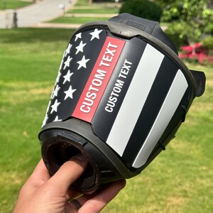May include: A black fire helmet front piece featuring a stylized American flag design with a thin red line. The decal includes the text CUSTOM TEXT printed twice in white and red lettering. The piece is constructed from molded plastic with the MSA logo.