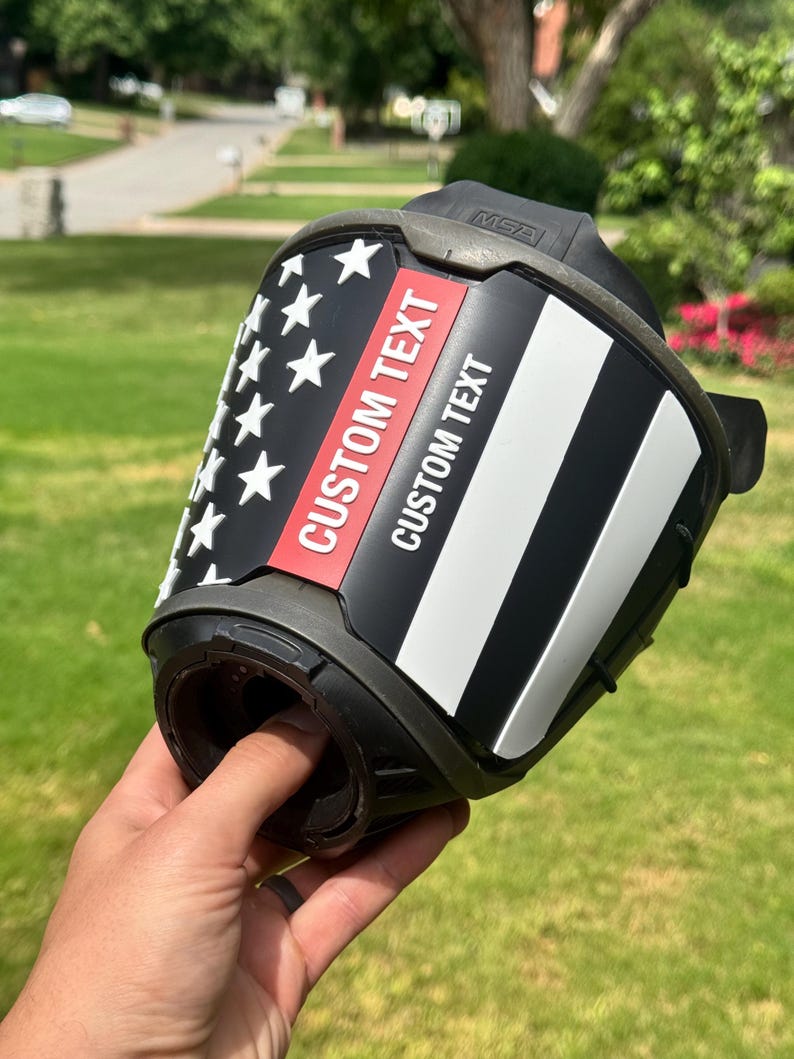 May include: A black fire helmet front piece featuring a stylized American flag design with a thin red line. The decal includes the text CUSTOM TEXT printed twice in white and red lettering. The piece is constructed from molded plastic with the MSA logo.