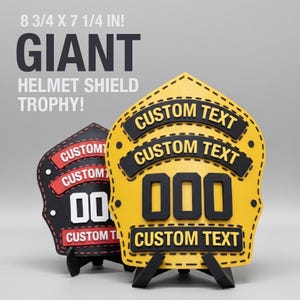 Puede incluir: Dos trofeos gigantes con forma de escudo de casco. Uno es amarillo con detalles negros y el otro es negro con detalles rojos. Cada uno tiene el texto "CUSTOM TEXT" y un número. Las dimensiones son de 22,2 x 18,4 cm.