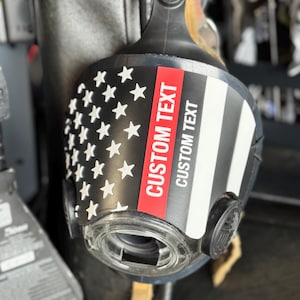 May include: A black respirator mask featuring a graphic of the American flag with a thin red line. The mask includes a clear circular filter port and a prominent red and white vertical stripe with the text CUSTOM TEXT printed twice.