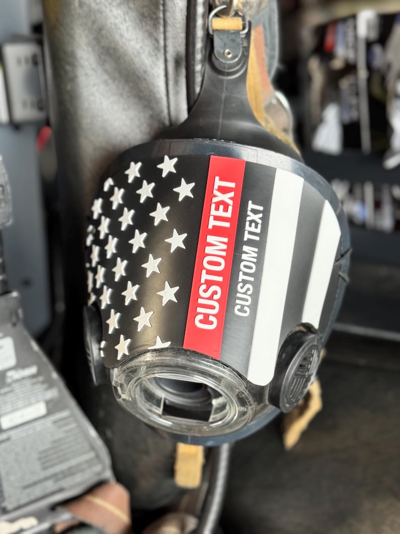 May include: A black respirator mask featuring a graphic of the American flag with a thin red line. The mask includes a clear circular filter port and a prominent red and white vertical stripe with the text CUSTOM TEXT printed twice.