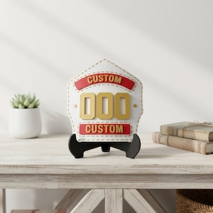 Centered image of a 3D printed firefighter helmet shield replica in a striking white and gold color scheme with red banner accents. The PLA display plaque features generic CUSTOM text and 000 numbers, showcased on a light wooden table next to a succulent and stacked books. Excellent as personalized fire department decor or a high-quality custom fireman gift.