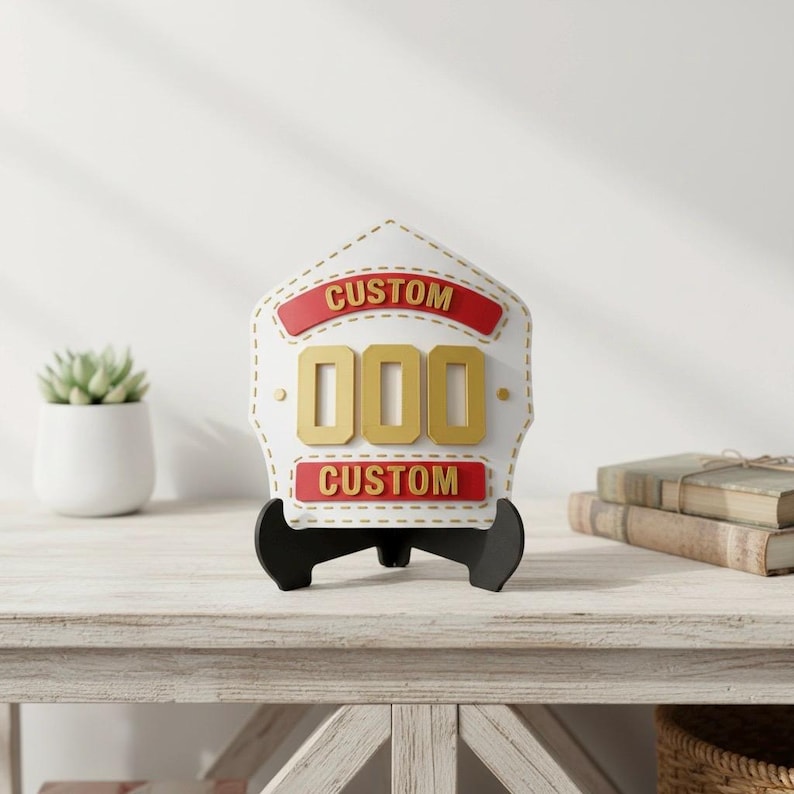 Centered image of a 3D printed firefighter helmet shield replica in a striking white and gold color scheme with red banner accents. The PLA display plaque features generic CUSTOM text and 000 numbers, showcased on a light wooden table next to a succulent and stacked books. Excellent as personalized fire department decor or a high-quality custom fireman gift.