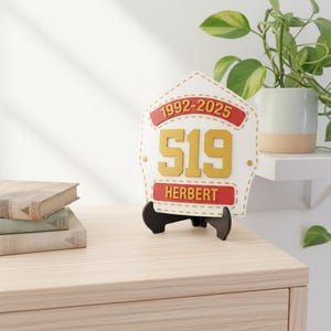 A white, red, and gold 3D Printed Fire Helmet Shield replica displayed on a black stand atop a wooden chest of drawers. The shield is customized with the years "1992-2025," the number "519," and the name "HERBERT," clearly marketing it as the ultimate personalized firefighter retirement gift. The image also includes a stack of vintage books and a potted plant, showing it as attractive home or office decor.
