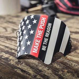 May include: A curved black, white, and red accessory featuring a stylized American flag design with white stars on the left and horizontal stripes on the right. The text "MAKE THE PUSH" and "BE THE REASON" is printed in white on the red and black sections.