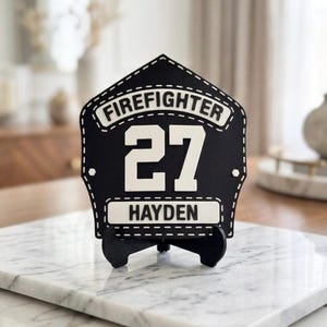 Pode incluir: Uma placa de bombeiro em forma de escudo, em preto e branco, com o texto FIREFIGHTER no topo, o n&uacute;mero 27 ao centro e o nome HAYDEN na parte inferior. A placa est&aacute; montada num pequeno suporte preto.