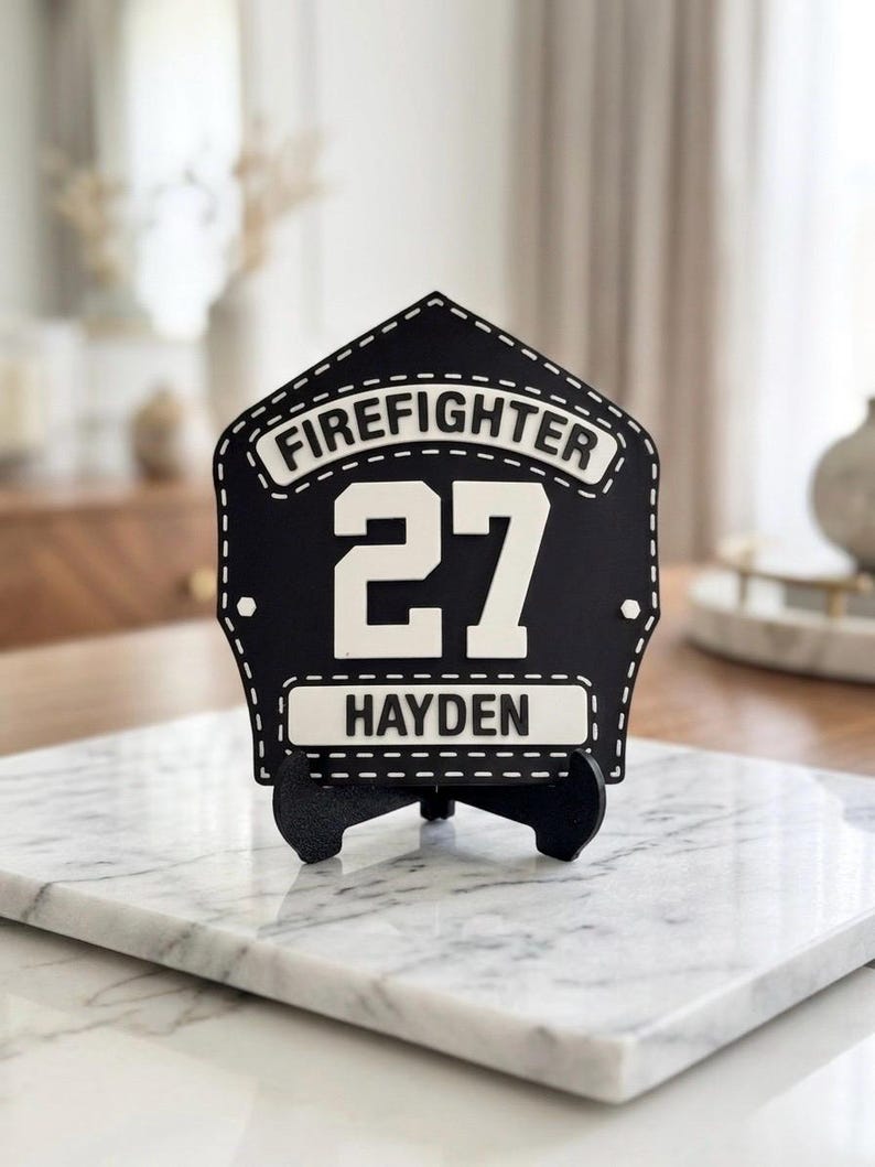 Pode incluir: Uma placa de bombeiro em forma de escudo, em preto e branco, com o texto FIREFIGHTER no topo, o n&uacute;mero 27 ao centro e o nome HAYDEN na parte inferior. A placa est&aacute; montada num pequeno suporte preto.