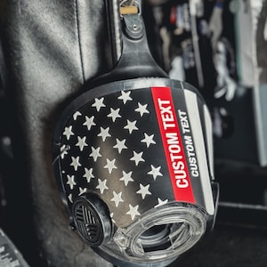 May include: A black respirator mask featuring a graphic of the American flag with white stars on a black field. A horizontal red stripe runs across the front, with the words CUSTOM TEXT printed in white on the red stripe and below it.
