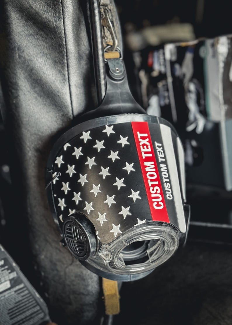 May include: A black respirator mask featuring a graphic of the American flag with white stars on a black field. A horizontal red stripe runs across the front, with the words CUSTOM TEXT printed in white on the red stripe and below it.