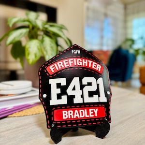 Extreme close-up of a highly personalized 3D printed firefighter shield in black and red. The shield features the text "FIREFIGHTER," Engine number E421, and the name "BRADLEY." The sharp detail of this custom PLA helmet replica is visible, set against a blurred indoor background, emphasizing it as a personal gift or desk plaque for an individual fireman.