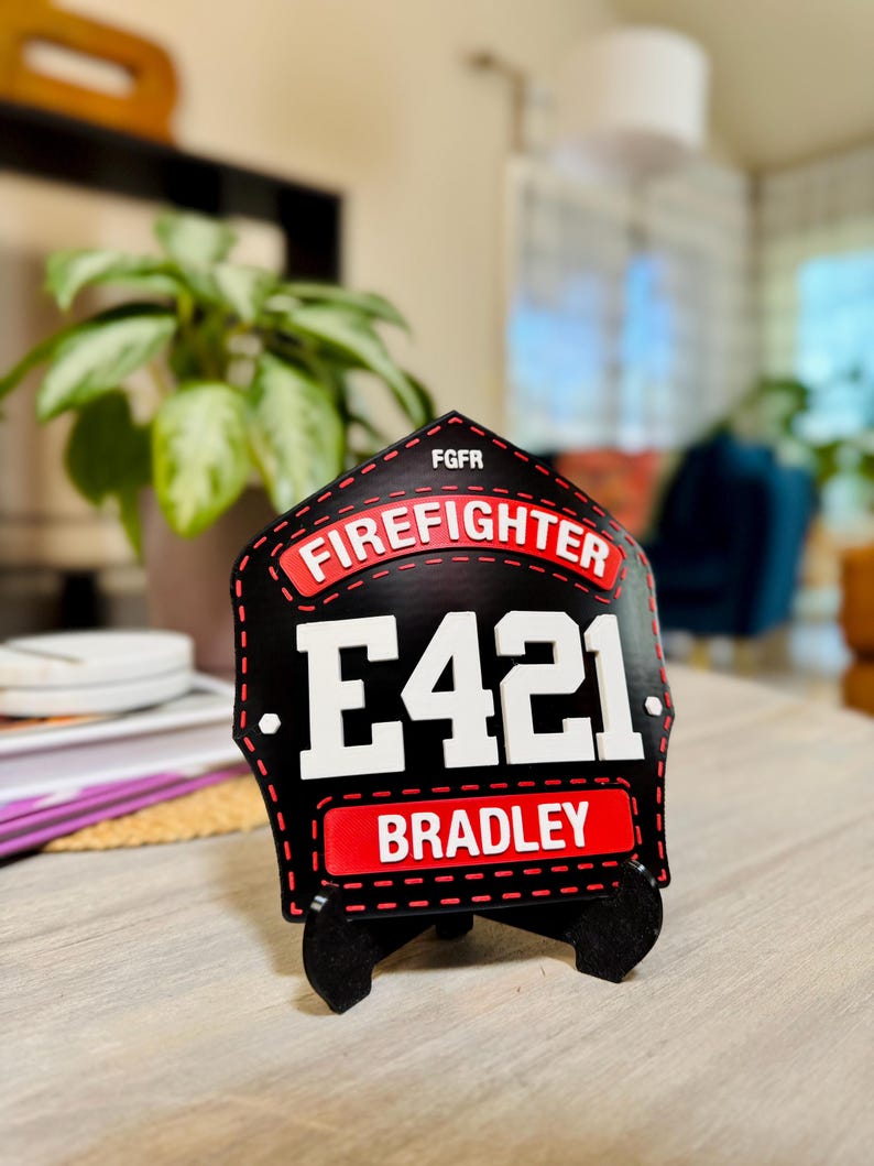 Extreme close-up of a highly personalized 3D printed firefighter shield in black and red. The shield features the text "FIREFIGHTER," Engine number E421, and the name "BRADLEY." The sharp detail of this custom PLA helmet replica is visible, set against a blurred indoor background, emphasizing it as a personal gift or desk plaque for an individual fireman.