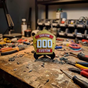 A 3D printed Fire Helmet Shield replica in yellow and red with large white customizable numbers ("000") stands centered on a messy wooden workbench surrounded by tools, wires, screwdrivers, and electronic components. This atmospheric photo highlights the handmade quality of the PLA fireman plaque and emphasizes the dedication behind this custom firefighter gift designed for a shop or garage.