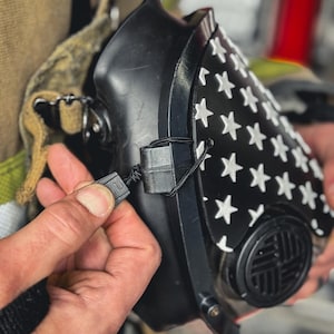May include: A black respirator mask featuring a white star pattern on the front panel. The mask includes a circular filter housing with a grid design and is secured with a thin black cord and a plastic adjustment toggle for a snug fit.