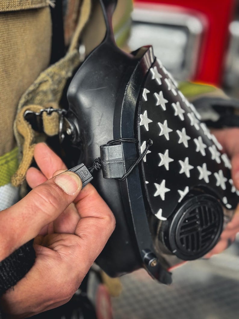 May include: A black respirator mask featuring a white star pattern on the front panel. The mask includes a circular filter housing with a grid design and is secured with a thin black cord and a plastic adjustment toggle for a snug fit.