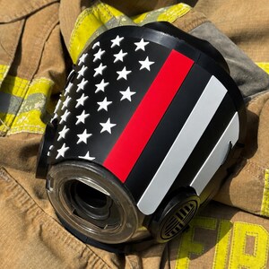 May include: A black respirator mask featuring a graphic of the American flag with a thin red line. The mask has a circular clear lens, side filter ports, and white star accents on the left side, with a red and white stripe pattern on the right.