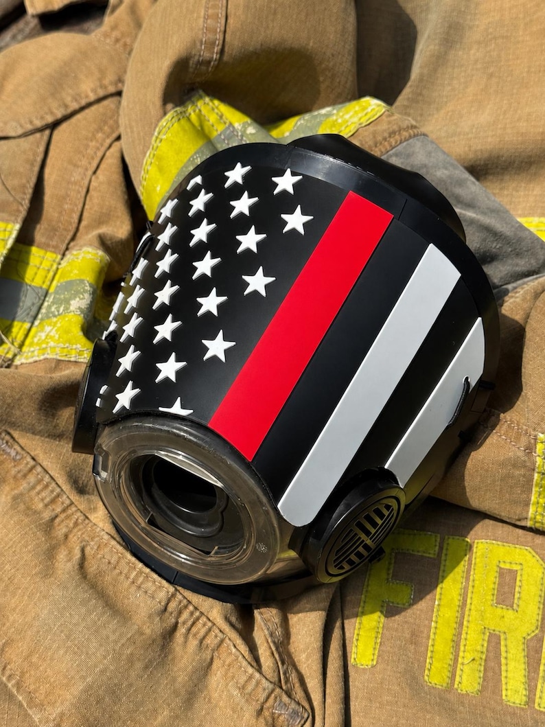 May include: A black respirator mask featuring a graphic of the American flag with a thin red line. The mask has a circular clear lens, side filter ports, and white star accents on the left side, with a red and white stripe pattern on the right.
