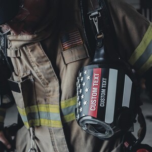 May include: A black firefighter helmet featuring a graphic of the American flag with a thin red line. The helmet displays the text CUSTOM TEXT in white and red lettering. It is worn with a tan protective jacket with yellow reflective stripes.