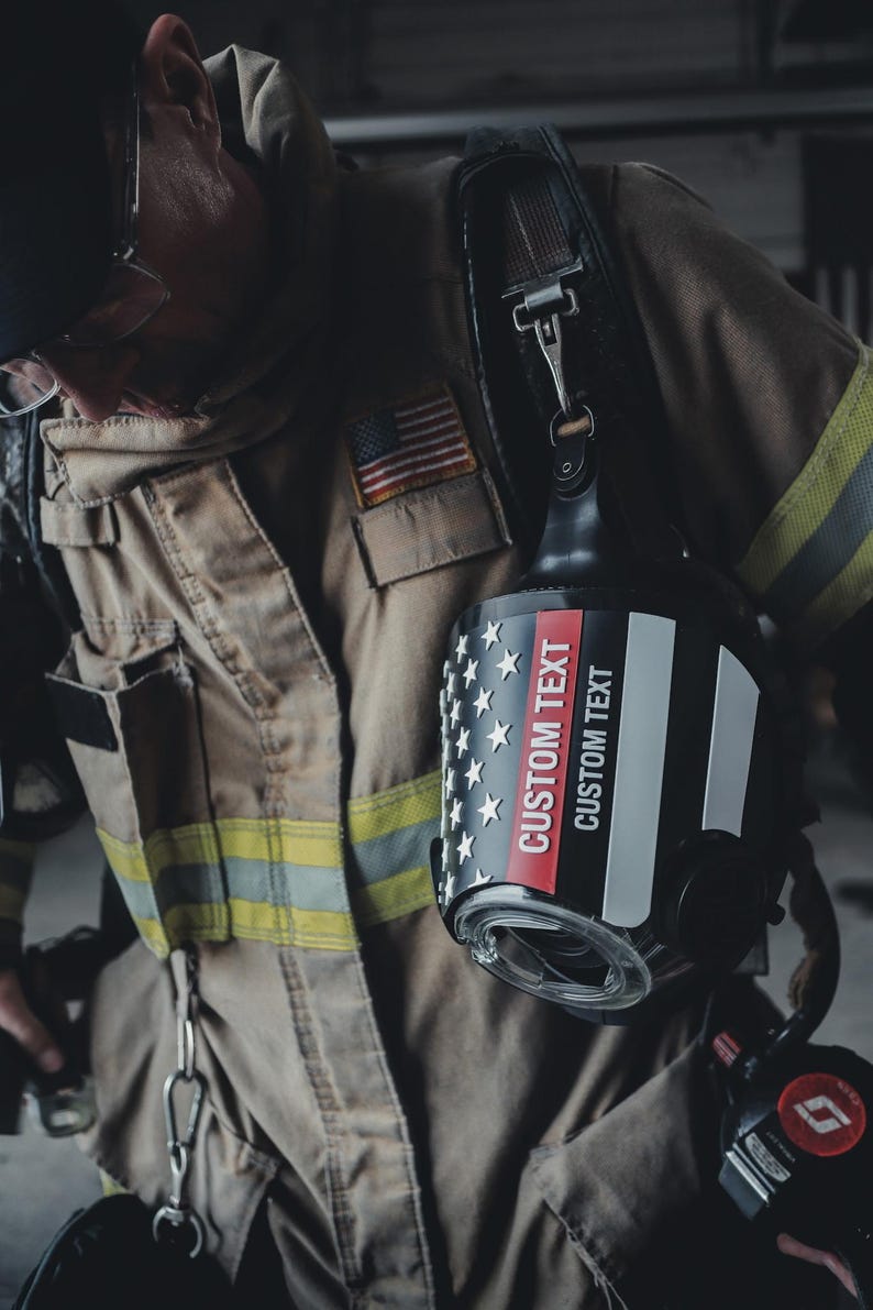 May include: A black firefighter helmet featuring a graphic of the American flag with a thin red line. The helmet displays the text CUSTOM TEXT in white and red lettering. It is worn with a tan protective jacket with yellow reflective stripes.