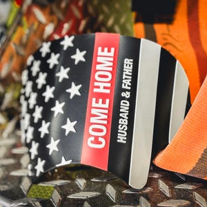 May include: A curved black, white, and red emblem featuring a stylized American flag design. The text "COME HOME" is printed in bold white letters on a vertical red stripe, with the words "HUSBAND & FATHER" in white below it on the black field.