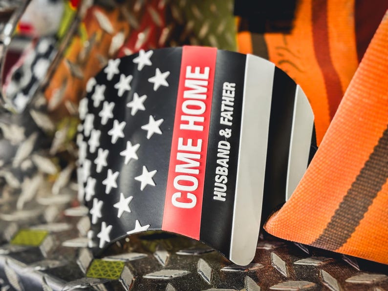 May include: A curved black, white, and red emblem featuring a stylized American flag design. The text "COME HOME" is printed in bold white letters on a vertical red stripe, with the words "HUSBAND & FATHER" in white below it on the black field.