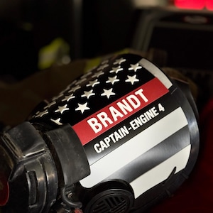 May include: A black firefighter helmet featuring a custom decal with a stylized American flag design, a thin red line, and white text that reads BRANDT and CAPTAIN-ENGINE 4. The helmet includes a black face piece and integrated mechanical components.