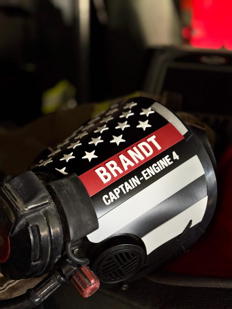 May include: A black firefighter helmet featuring a custom decal with a stylized American flag design, a thin red line, and white text that reads BRANDT and CAPTAIN-ENGINE 4. The helmet includes a black face piece and integrated mechanical components.