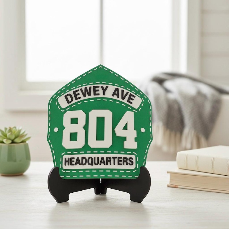 Close-up of a custom 3D Printed Fire Helmet Shield in a unique green and white color scheme, displayed on a black easel stand. The PLA replica features white text reading "DEWEY AVE" and "HEADQUARTERS" with the large number "804." This image showcases a personalized fire station sign, an excellent firehouse decor gift, or a custom fireman's plaque for a station chief's desk.