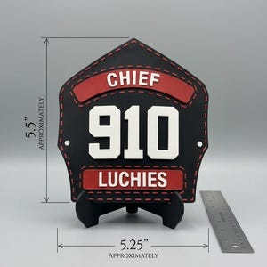 Pode incluir: Uma placa frontal de capacete de bombeiro em forma de escudo, preta e vermelha, com o texto CHIEF, 910 e LUCHIES. O item mede aproximadamente 13,97 cm de altura e 13,34 cm de largura, conforme indicado pela escala vis&iacute;vel.