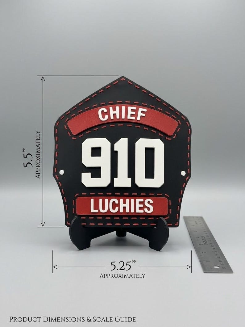 Pode incluir: Uma placa frontal de capacete de bombeiro em forma de escudo, preta e vermelha, com o texto CHIEF, 910 e LUCHIES. O item mede aproximadamente 13,97 cm de altura e 13,34 cm de largura, conforme indicado pela escala vis&iacute;vel.