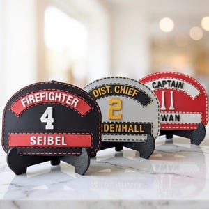 Puede incluir: Tres placas frontales semicirculares para cascos de bombero sobre soportes negros. La primera es negra con detalles rojos y dice FIREFIGHTER 4 SEIBEL. La segunda es blanca con detalles negros y dice DIST. CHIEF 2 MIDENHALL. La tercera es roja con detalles blancos y dice CAPTAIN 11 SWAN.