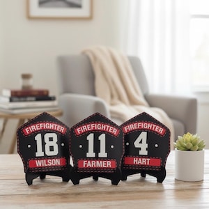 Three customized black firefighter helmet shields displayed side-by-side on a wooden surface in a soft-focus indoor setting. The shields feature white numbers (18, 11, 4) and names (WILSON, FARMER, HART) beneath the "FIREFIGHTER" banner, highlighting the multiple design and personalization options available for fire crew gifts or fire station retirement plaques.