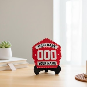 A close-up, centered image of a 3D printed Firefighter Helmet Shield replica in red, featuring white customizable text areas for "YOUR RANK" and "YOUR NAME" with bold white numbers ("000"). The PLA display shield is presented on a wooden desk with a black stand, making it an excellent custom fireman retirement gift, desk plaque, or firefighter promotion present.