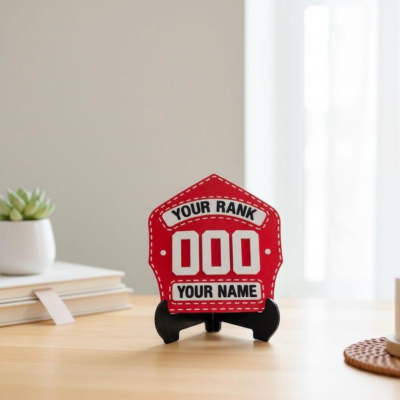 A close-up, centered image of a 3D printed Firefighter Helmet Shield replica in red, featuring white customizable text areas for "YOUR RANK" and "YOUR NAME" with bold white numbers ("000"). The PLA display shield is presented on a wooden desk with a black stand, making it an excellent custom fireman retirement gift, desk plaque, or firefighter promotion present.