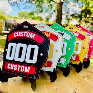 A line of multiple colored 3D printed firefighter helmet shields standing on a wooden slab outdoors. The shields are shown in a row from black to white/yellow, green, and red, showcasing the full spectrum of custom color options. This bright, outdoor shot emphasizes the PLA replica's durability and wide range of custom firehouse decor available.