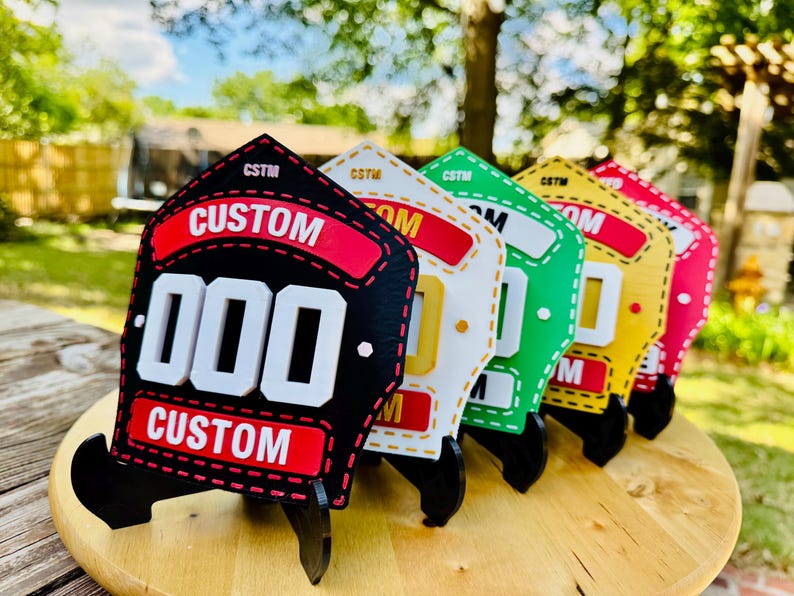 A line of multiple colored 3D printed firefighter helmet shields standing on a wooden slab outdoors. The shields are shown in a row from black to white/yellow, green, and red, showcasing the full spectrum of custom color options. This bright, outdoor shot emphasizes the PLA replica's durability and wide range of custom firehouse decor available.