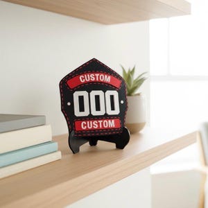 Close-up of a single 3D printed firefighter helmet shield replica, colored black and red with large white customizable numbers ("000") and "CUSTOM" text. The PLA shield sits on a black display stand on a light wood floating shelf next to a stack of books and a small potted succulent, showcasing it as firehouse desk decor or a personalized fireman gift for an office.