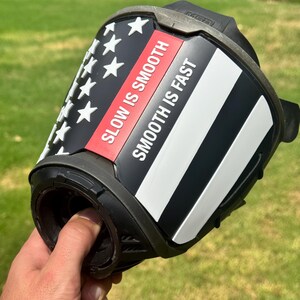 May include: A black helmet accessory featuring a stylized American flag design with white stars and stripes. A red horizontal stripe contains the white text SLOW IS SMOOTH, while a white stripe below it features the text SMOOTH IS FAST in black.