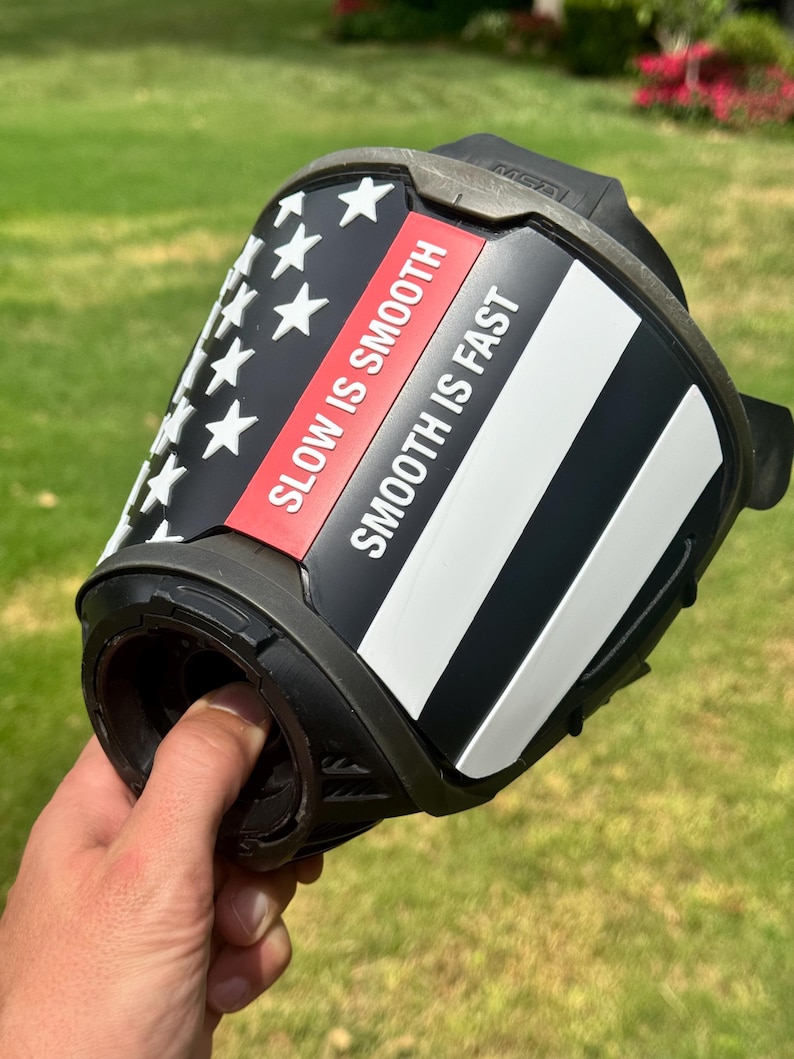 May include: A black helmet accessory featuring a stylized American flag design with white stars and stripes. A red horizontal stripe contains the white text SLOW IS SMOOTH, while a white stripe below it features the text SMOOTH IS FAST in black.