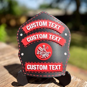 Puede incluir: Un casco de bombero negro con detalles rojos y el texto "CUSTOM TEXT" en tres pancartas rojas. Un círculo rojo con un emblema de bombero está centrado en el casco. El casco se muestra sobre una superficie de madera.